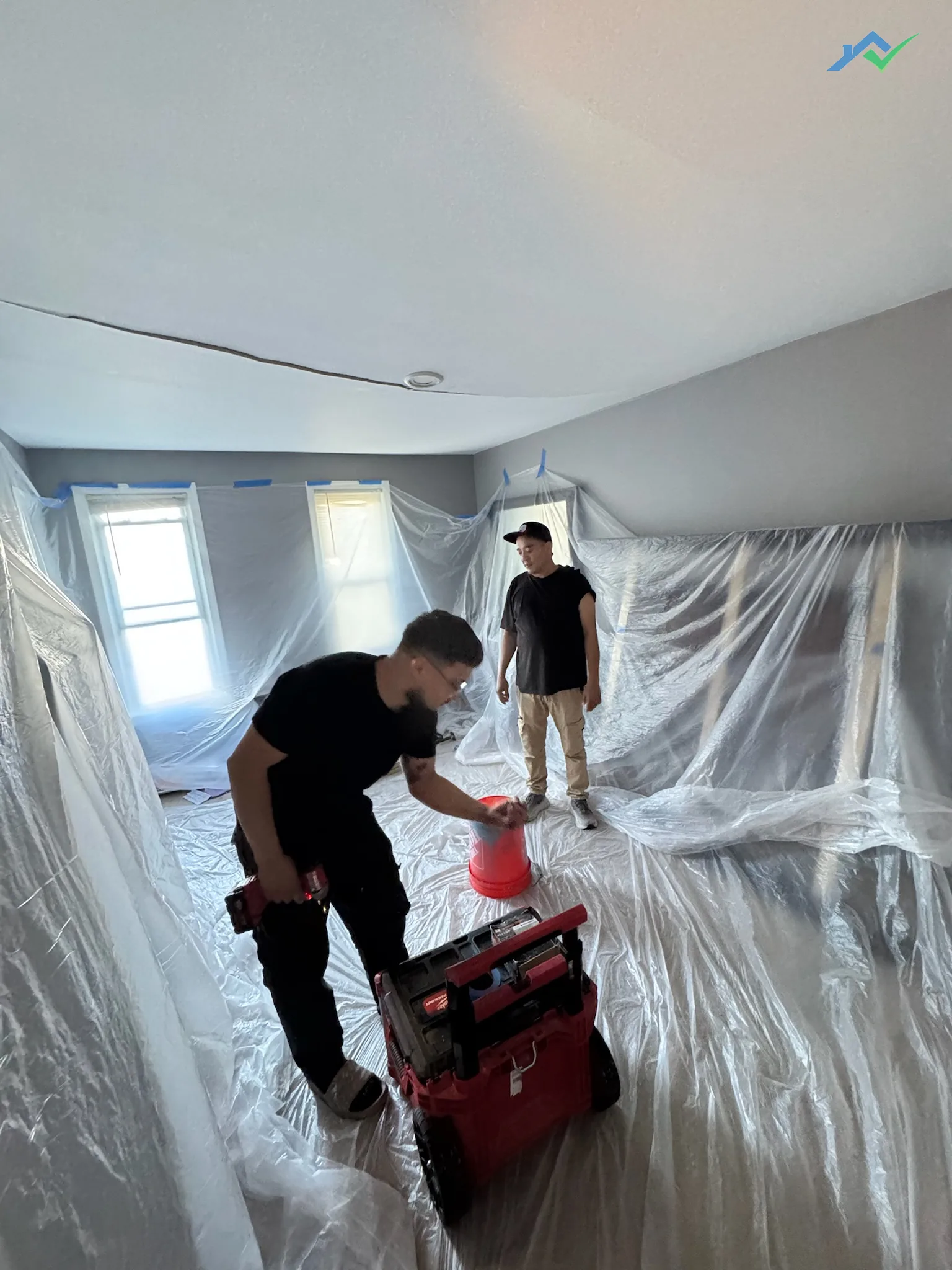 The Burke bedroom fully wrapped in plastic sheeting and blue painter's tape before ceiling demolition, with two crew members setting up.