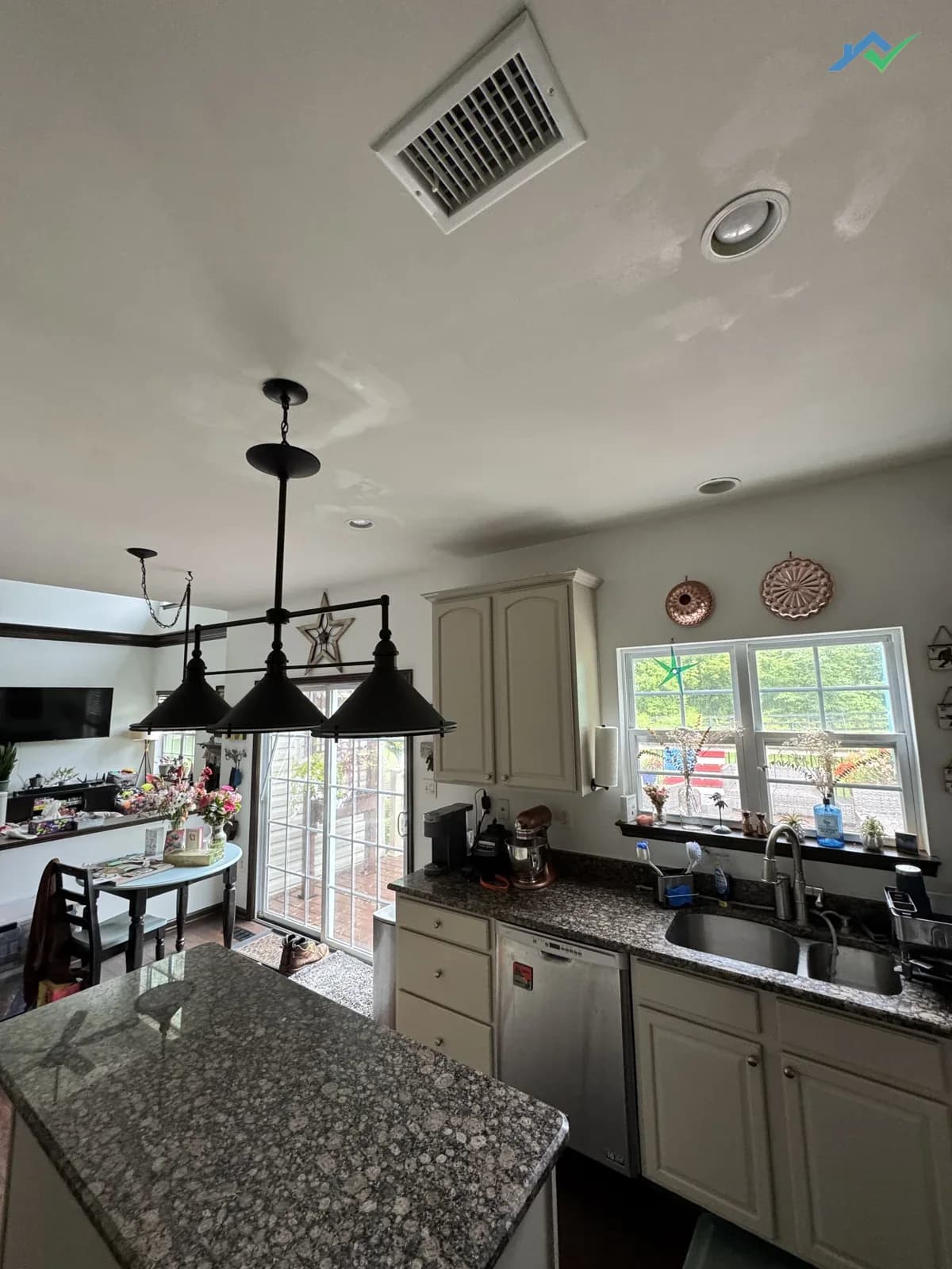 Same Purcellville, VA kitchen ceiling after repair — the water stain trail around the louvered HVAC vent and recessed light is completely gone, the ceiling reads uniformly white above the cream cabinets and pendant lights with no ghosting or bleed-through.