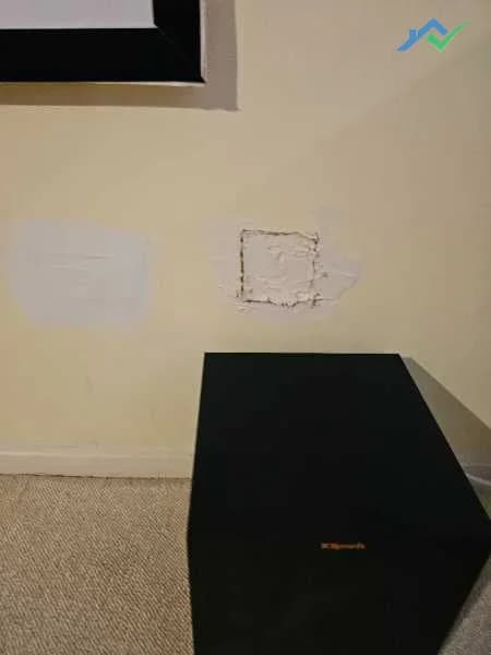 Before shot of a basement wall in this Purcellville, VA home — two rough previous patch attempts side by side on the yellow-tan wall next to the projector-screen home theater setup, one lightly skimmed and one still showing the raw plug, neither ever finished properly.