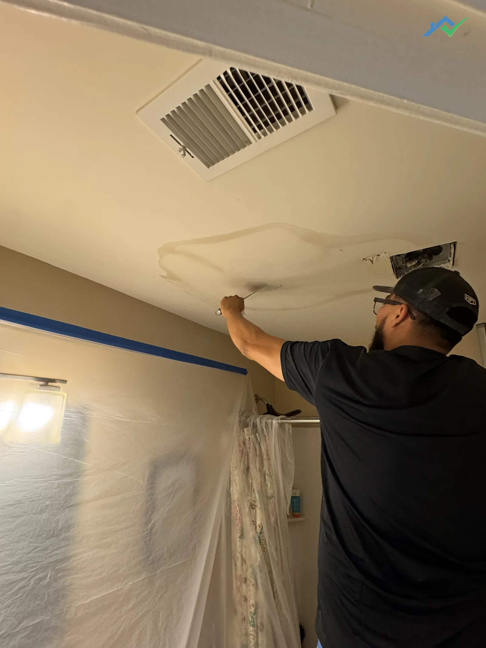 Eduardo on a step ladder in the Purcellville, VA home repairing nail pops and preparing the previously water-stained ceiling with stain-blocking primer, with blue painter's tape and plastic containment sheeting protecting the walls below.