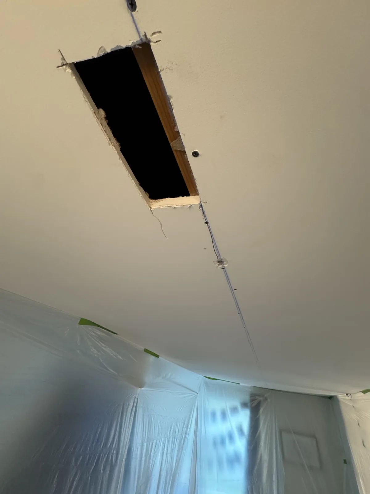 Access cutout opened in a Herndon, VA ceiling exposing the joist so HouseWorks could inspect the framing and re-secure the drywall panel — plastic containment is already in place below to protect the room.