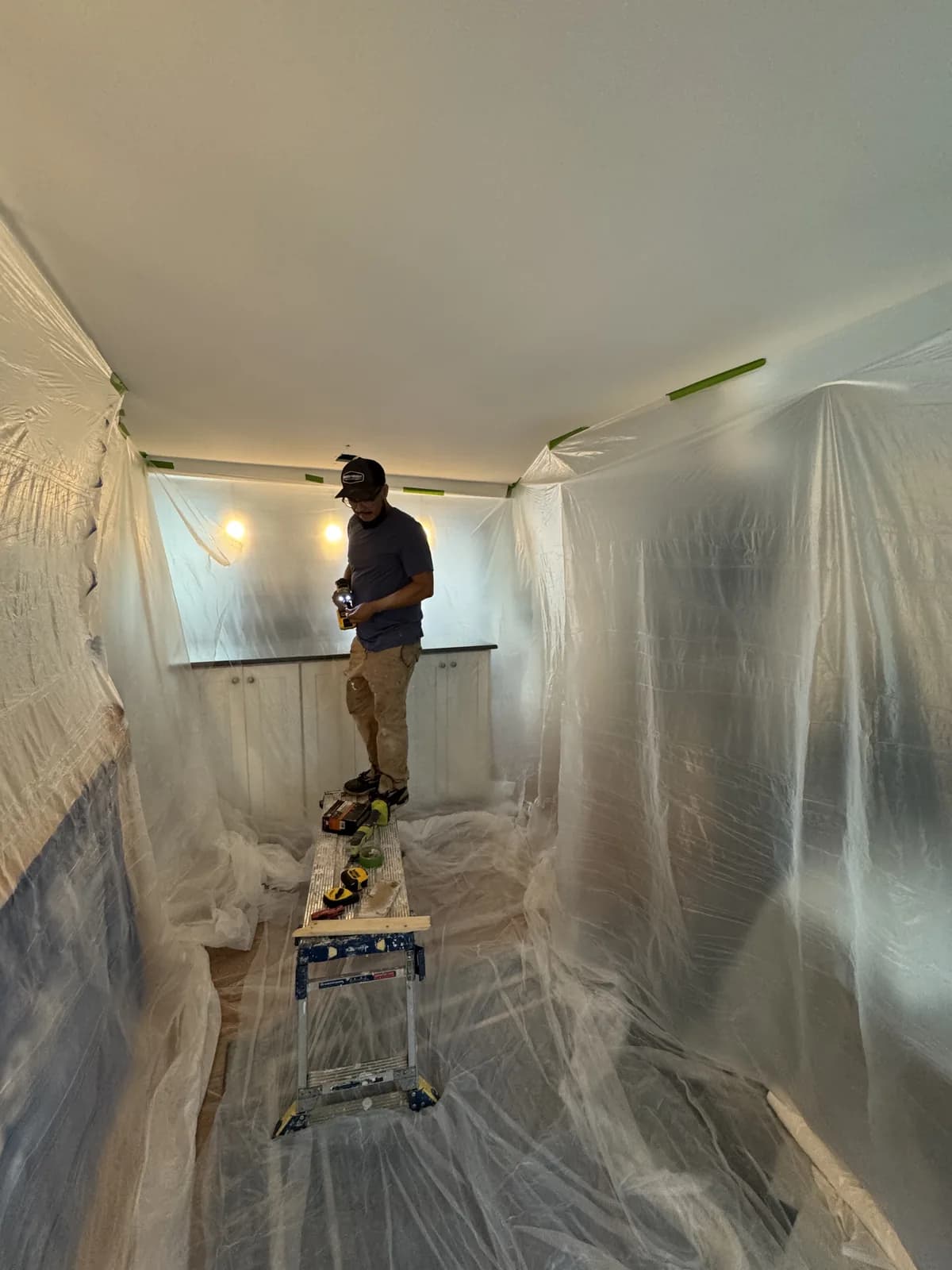 HouseWorks tech Eduardo on a step ladder in a Herndon, VA home, drill in hand, working on the ceiling with the entire room wrapped in clear plastic containment to protect walls and floors from dust.